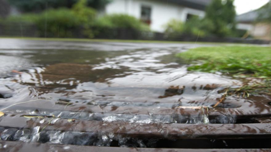 Regenwasser fließt in einen Straßengulli, im Hintergrund unscharf ein weißes Haus